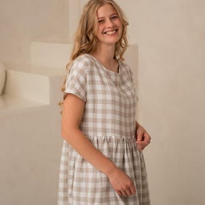 May include: A beige and white checkered dress with short sleeves. The dress has a round neckline and a gathered waist. The fabric appears to be linen or a similar lightweight material. The dress is knee-length and suitable for casual wear.