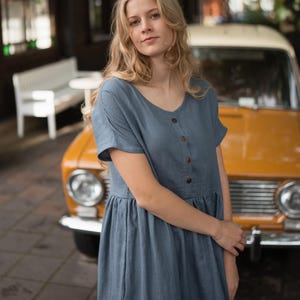 May include: A woman wearing a blue dress with short sleeves and wooden buttons. The dress has a simple, flowing design. The background includes a vintage yellow car and a white bench, suggesting a casual, outdoor setting.