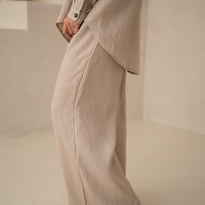 May include: Beige linen pants and a matching long-sleeved shirt. The outfit has a relaxed fit and is made of a lightweight fabric. The pants are wide-legged, and the shirt has buttoned cuffs. The outfit is suitable for casual wear.