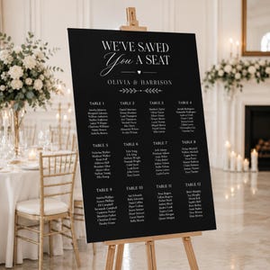 May include: A black wedding seating chart sign on a wooden easel with white text that reads WE'VE SAVED YOU A SEAT, OLIVIA & HARRISON, followed by a list of guests organized by twelve tables. The sign features a minimalist design with white font.