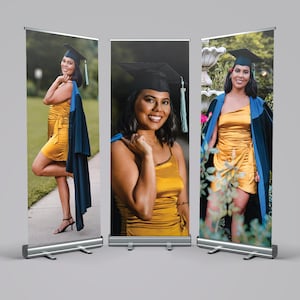 May include: Three graduation banners featuring a woman in a gold dress, black cap, and blue graduation gown. The banners are displayed on silver retractable stands, showcasing a celebratory theme. The woman is smiling in each image.