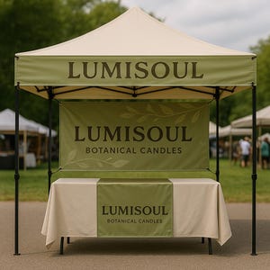 May include: A pop-up tent with a cream-colored canopy and a green banner displaying the text "LUMISOUL BOTANICAL CANDLES." The tent has a table covered with a cream tablecloth and a green runner. The tent is set up outdoors.