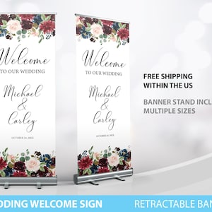 May include: Two retractable wedding welcome signs. Each sign features the text "Welcome to our wedding" above the names "Michael & Carley" and the date "October 24, 20XX". The signs have a floral design with burgundy, navy, and blush-colored flowers.