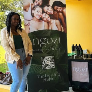 May include: A woman stands next to a banner and table display. The banner features a group of smiling women and the text "ngozi" and "The blessing of skin." The table displays several dark bottles. The woman wears a gold sequined jacket, green top, and blue jeans.