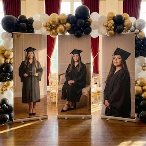 May include: Three graduation banners featuring a woman in a black cap and gown. The banners are set up in front of a balloon arch with black, gold, silver, and white balloons. The background includes red curtains and a window.