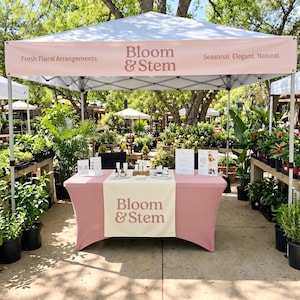 May include: A white pop-up canopy tent with a pink valance featuring the text Bloom & Stem, Fresh Floral Arrangements, and Seasonal. Elegant. Natural. A pink table cover with a cream runner displays the text Bloom & Stem and various bottles.