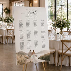 May include: A white wedding seating chart sign on a gold easel featuring the text Please find your seat, Lauren & Michael, June 21, 2025, a list of twelve tables with guest names, and a photograph of a couple walking on a path toward mountains.