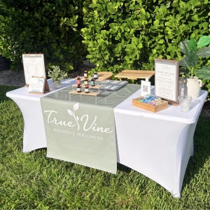 May include: A white table covered with a sage green runner featuring the white text True Vine Women's Wellness. The display includes a tiered wooden stand with small bottles, a wooden box with packets, two framed signs, and a potted plant.