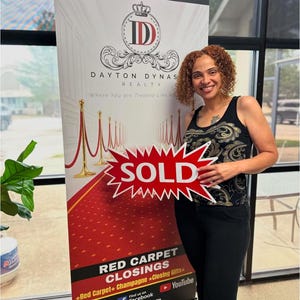 May include: A woman stands next to a banner for Dayton Dynas Realty. The banner features a red carpet design and the text "Where You are Treated Like Royalty." She holds a red sign that says "SOLD."