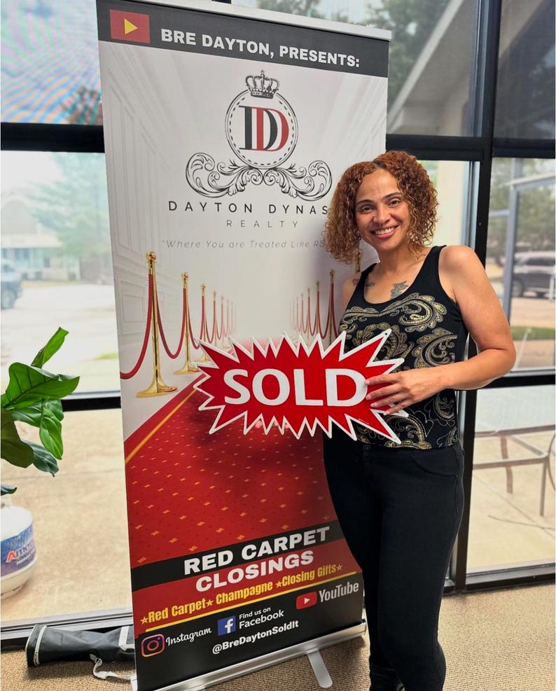 May include: A woman stands next to a banner for Dayton Dynas Realty. The banner features a red carpet design and the text "Where You are Treated Like Royalty." She holds a red sign that says "SOLD."