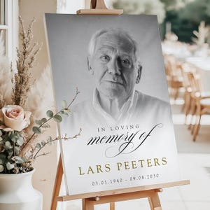 May include: A memorial sign on an easel features a black and white portrait of an older man. The sign reads "In Loving Memory of Lars Peeters" with dates 03.01.1940 - 09.08.2030 in gold. The sign is displayed indoors.