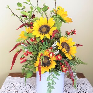May include: A white metal watering can filled with a floral arrangement of sunflowers, red berries, and green foliage. The arrangement is on a white crocheted tablecloth.