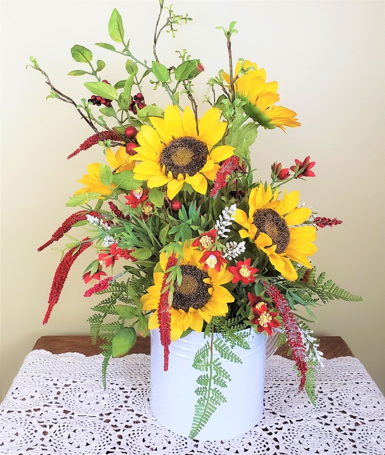 May include: A white metal watering can filled with a floral arrangement of sunflowers, red berries, and green foliage. The arrangement is on a white crocheted tablecloth.