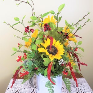 May include: A white watering can with a black and white striped handle filled with a floral arrangement of sunflowers, red berries, and green foliage.