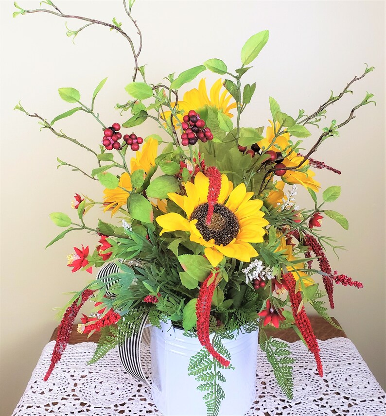 May include: A white watering can with a black and white striped handle filled with a floral arrangement of sunflowers, red berries, and green foliage.