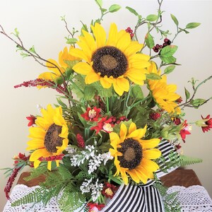 May include: A bouquet of sunflowers in a white vase with black and white striped ribbon. The sunflowers are yellow with dark brown centers. The vase is decorated with red berries and green leaves.