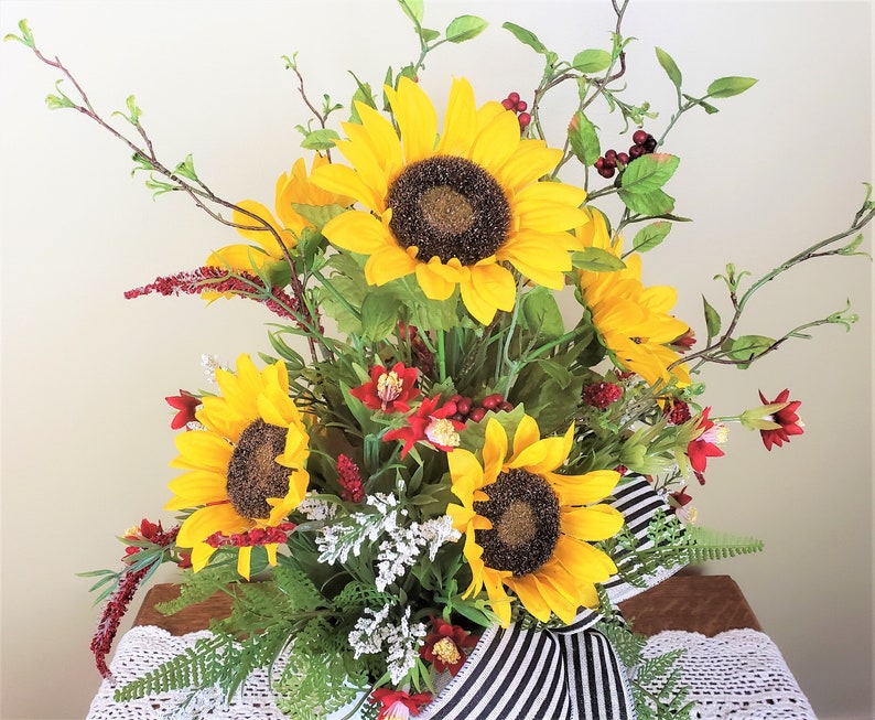 May include: A bouquet of sunflowers in a white vase with black and white striped ribbon. The sunflowers are yellow with dark brown centers. The vase is decorated with red berries and green leaves.
