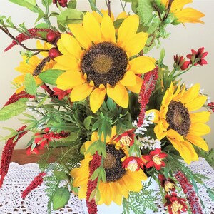 May include: A bouquet of artificial sunflowers and red flowers in a white vase. The sunflowers have yellow petals and dark brown centers. The red flowers are small and have a delicate texture.