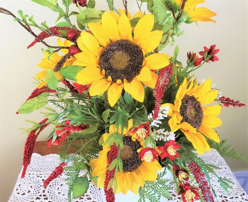 May include: A bouquet of artificial sunflowers and red flowers in a white vase. The sunflowers have yellow petals and dark brown centers. The red flowers are small and have a delicate texture.