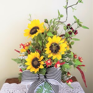 May include: A bouquet of sunflowers and red berries arranged in a white pitcher with a black and white striped bow. The sunflowers are yellow with dark centers and the berries are small and round.