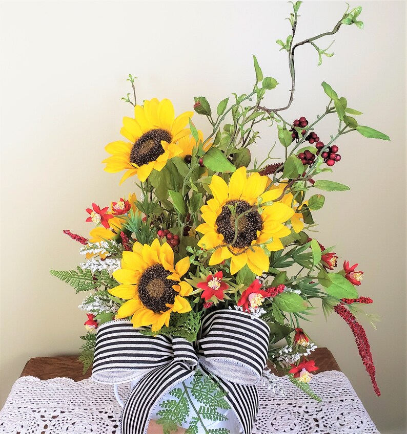 May include: A bouquet of sunflowers and red berries arranged in a white pitcher with a black and white striped bow. The sunflowers are yellow with dark centers and the berries are small and round.