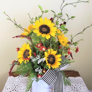 May include: A white metal bucket filled with a floral arrangement featuring yellow sunflowers, red berries, and green foliage. The bucket is decorated with a black and white striped ribbon.