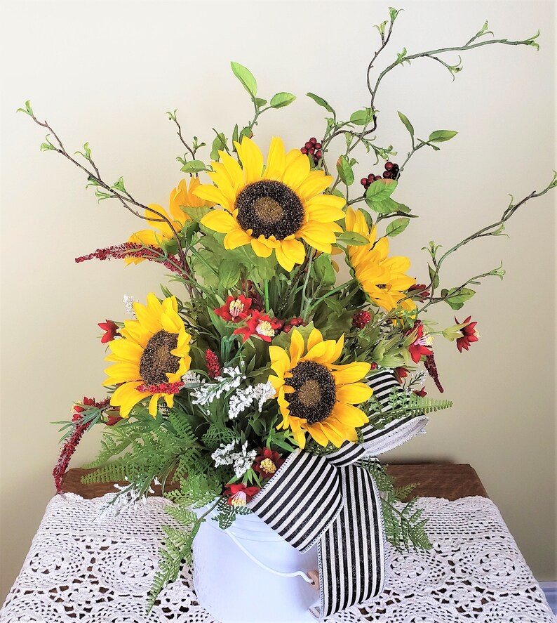 May include: A white metal bucket filled with a floral arrangement featuring yellow sunflowers, red berries, and green foliage. The bucket is decorated with a black and white striped ribbon.
