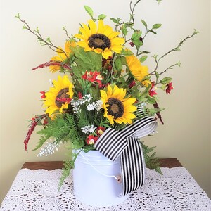 May include: A white metal pitcher with a black and white striped ribbon, filled with a floral arrangement of sunflowers, red and white flowers, and green foliage.
