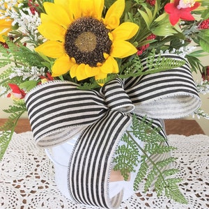 May include: A white ceramic pot with a black and white striped ribbon bow. The pot is filled with a floral arrangement of sunflowers, red flowers, and greenery.