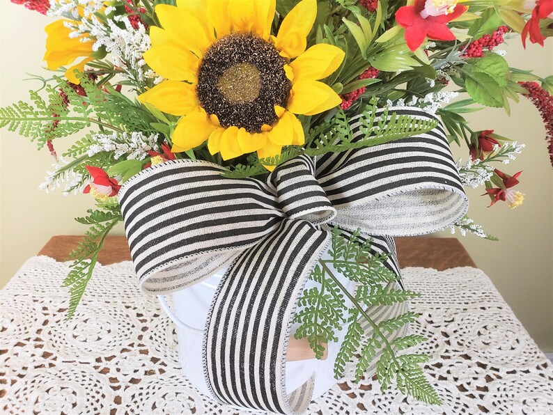 May include: A white ceramic pot with a black and white striped ribbon bow. The pot is filled with a floral arrangement of sunflowers, red flowers, and greenery.