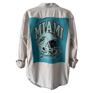 May include: A long-sleeved button-down shirt with a light beige and white striped pattern. The back features a turquoise panel with a Miami Dolphins football helmet graphic and the word "MIAMI" in white lettering. The shirt has rolled-up sleeves.