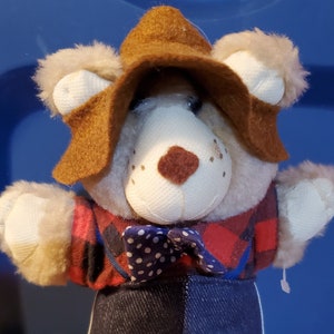 May include: A plush bear doll wearing a brown hat, a red and black plaid shirt, a blue bow tie with white polka dots, and denim pants. The bear has tan boots and fluffy ears. The doll is held by a hand.