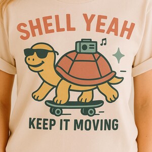 Könnte beinhalten: Cremefarbenes T-Shirt mit einer Cartoon-Schildkröte, die eine Sonnenbrille trägt und Skateboard fährt. Das Shirt zeigt den Text "SHELL YEAH" über der Schildkröte und "KEEP IT MOVING" darunter.