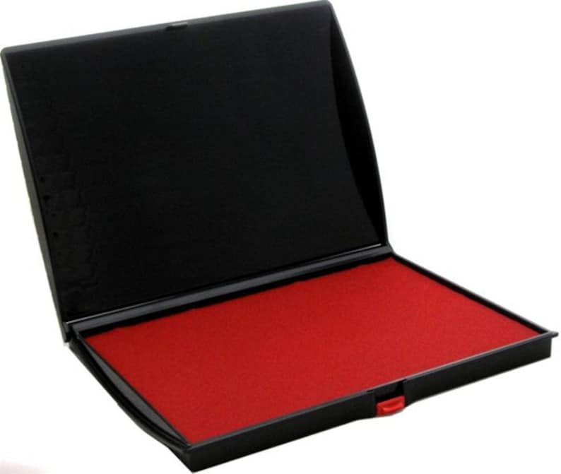 Puede incluir: Una almohadilla de tinta negra con una almohadilla de tinta roja en su interior. La almohadilla de tinta est&aacute; abierta y la almohadilla de tinta roja es visible.