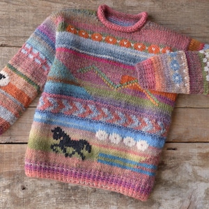 Op de afbeelding: Een kleurrijke gebreide trui met een geometrisch patroon met een paard en een schaap. De trui is gemaakt van verschillende kleuren, waaronder roze, blauw, groen, oranje en bruin.
