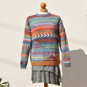 Könnte beinhalten: Ein bunter Strickpullover mit geometrischem Muster in Blautönen, Grün, Orange und Lila. Der Pullover hat einen Rundhalsausschnitt und lange Ärmel.