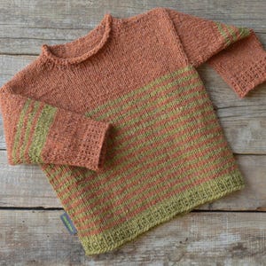 Könnte beinhalten: Ein gestrickter Pullover in Pfirsich- und Olivgrüntönen. Der Pullover hat horizontale olivgrüne Streifen über der unteren Hälfte und an den Ärmeln. Der Halsausschnitt und die Manschetten sind einfarbig pfirsichfarben. Das Kleidungsstück wird auf einer Holzoberfläche präsentiert.