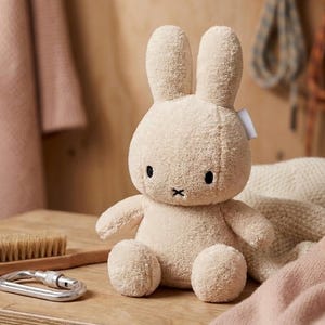 May include: A beige bunny plush toy with black eyes and a stitched black "X" mouth. The toy sits on a wooden surface with a wooden brush and a silver carabiner. A soft blanket and pink towel are in the background. The bunny has long ears.