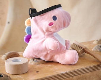 May include: A soft, light pink plush dinosaur chalk bag featuring a black rim, a white belly, and a row of small, colourful spikes in yellow, blue, and purple along its back. The dinosaur has embroidered dark blue eyes and pink cheeks.