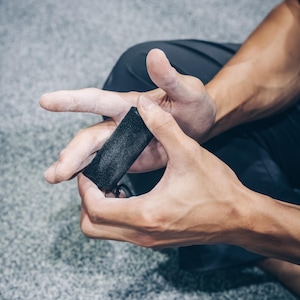 May include: A black climbing chalk block is held between two hands. The hands are covered in white chalk. The person is wearing dark gray pants. The background is a light gray carpet.
