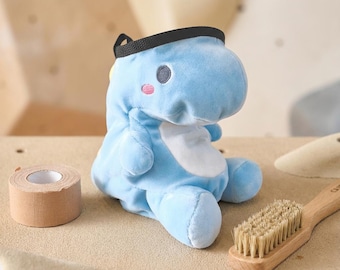 May include: A light blue plush dinosaur-shaped chalk bag with a white belly, dark grey eyes, and pink cheeks. The bag features a black rim opening at the top. Beside it sits a roll of tan athletic tape and a wooden brush with light bristles.