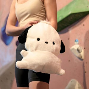 Puede incluir: Un juguete de peluche blanco con forma de perro con orejas y ojos negros. El juguete está siendo sostenido por una persona que lleva pantalones cortos negros y una camiseta de tirantes beige.