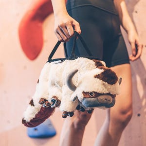 Może przedstawiać: Pluszowa zabawka w kształcie jaka, w kolorze białym i brązowym, z czarnym paskiem do noszenia. Zabawkę trzyma osoba w czarnych spodenkach.