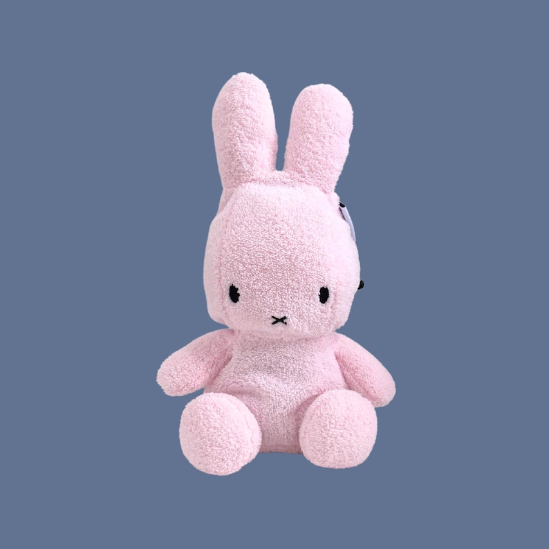 May include: A light pink plush rabbit toy with a soft, textured fleece-like material. The rabbit features two long upright ears, two small black oval eyes, and a simple black cross-stitched mouth. It has a rounded head and body.