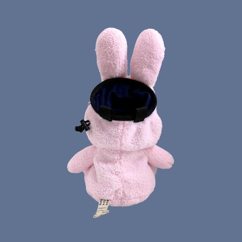 May include: A light pink plush rabbit-shaped bag with long ears and a textured fabric exterior. The bag features a circular black opening at the top with a drawstring closure, a black plastic toggle, and a small white fabric tag at the base.