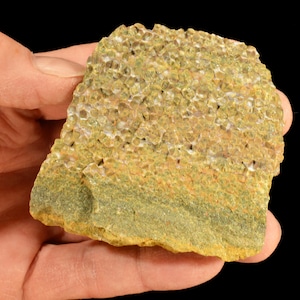 May include: A hand holding a piece of rough, textured rock. The rock has a layered appearance with shades of green, yellow, and orange. The top layer has a porous, honeycomb-like texture.