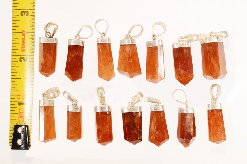 May include: A collection of orange-brown gemstone pendants with silver-colored metal caps and loops. The pendants are various shapes, including rectangular and faceted, and are approximately 1 to 2 inches in length. The background is white.