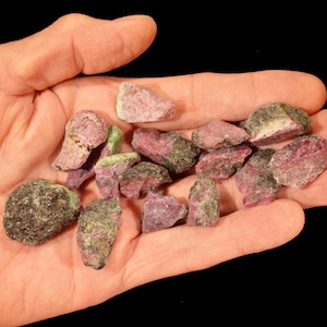 May include: A handful of rough-cut gemstones, primarily in shades of pink and green, are displayed against a black background. The stones vary in size and shape, showcasing a natural, unpolished aesthetic. The gemstones are held in an open hand.