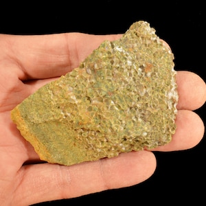 May include: A piece of rough, textured rock with a mix of green, yellow, and white colors. The rock has a porous surface and is held in a hand against a black background. The rock appears to be a natural mineral specimen.