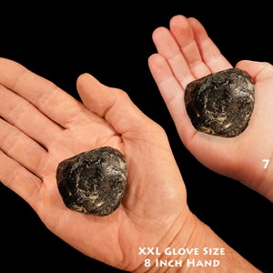 May include: Two hands holding a large, dark, round, smooth stone. The stone is about the size of a baseball. The hand on the left is an adult hand, wearing an XXL glove size. The hand on the right is a child's hand, belonging to a 7-year-old.